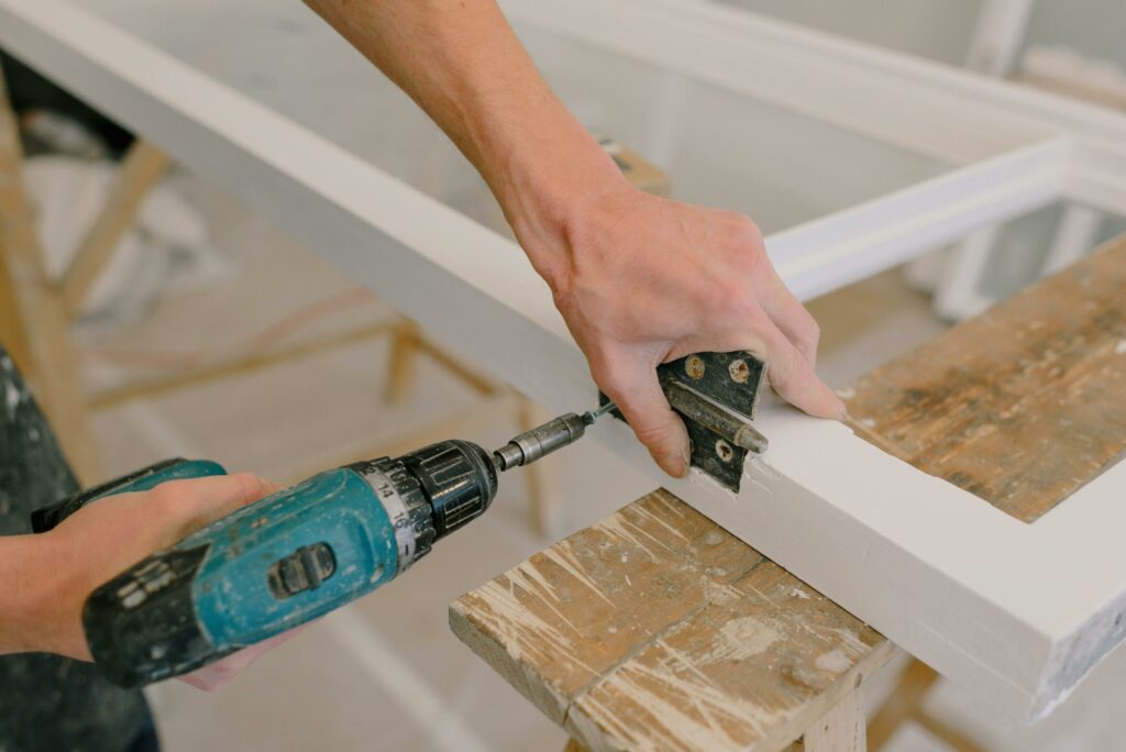 pexels photo 5691502 5691502 Unrecognizable professional male master using cordless screwdriver to install metal hinge on wooden window in light room during repair works
